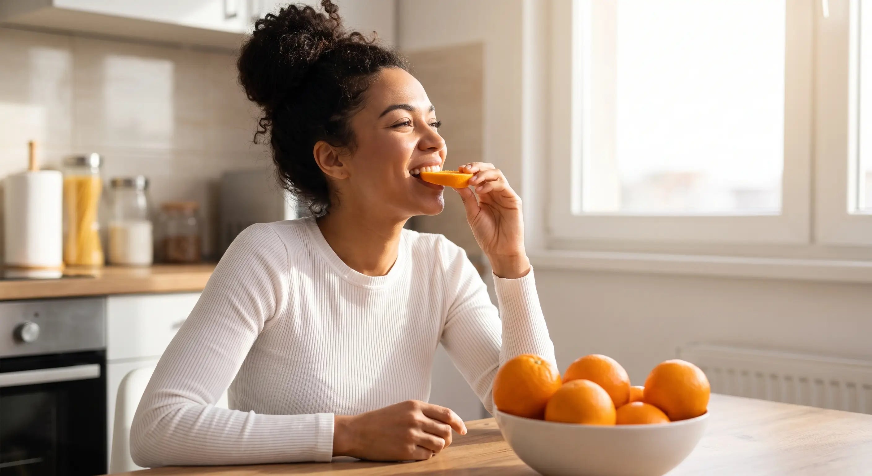 Une femme souriante assise dans une cuisine lumineuse suce un quartier d'orange fraîche, un bol rempli d'oranges posé sur la table en bois devant elle.